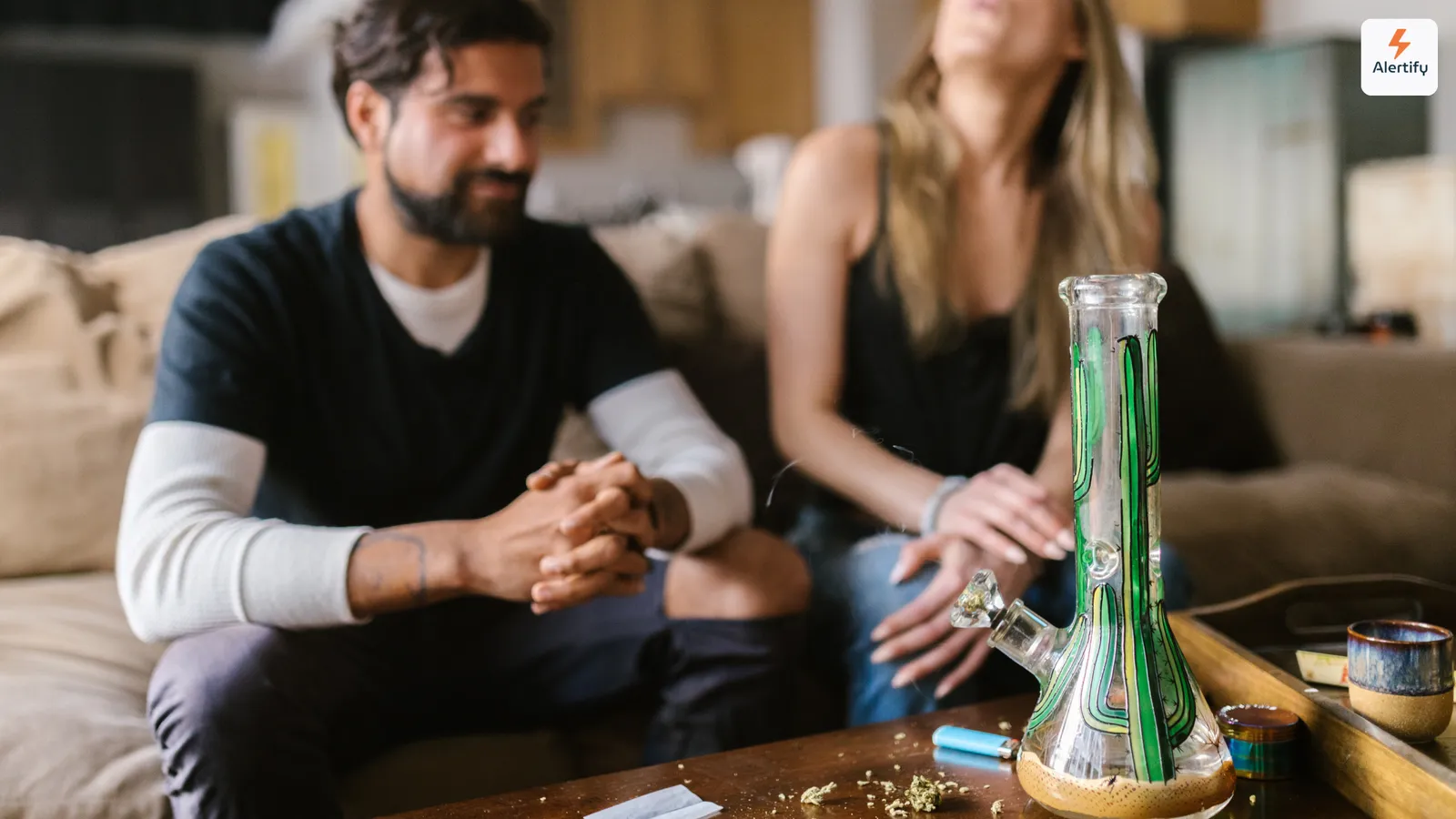 Due ospiti seduti su un divano nel soggiorno di un affitto, con un bong di vetro, un accendino e un grinder sul tavolino davanti a loro.