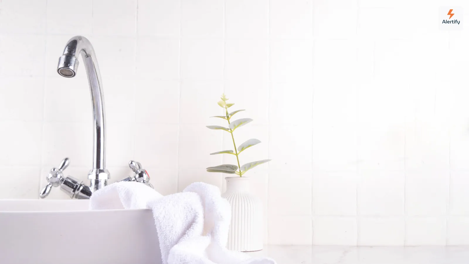 Un bagno luminoso rivestito di piastrelle bianche, con lavabo, un asciugamano piegato e una piccola pianta in vaso — il tipo di stanza umida dove la muffa attecchisce per prima.